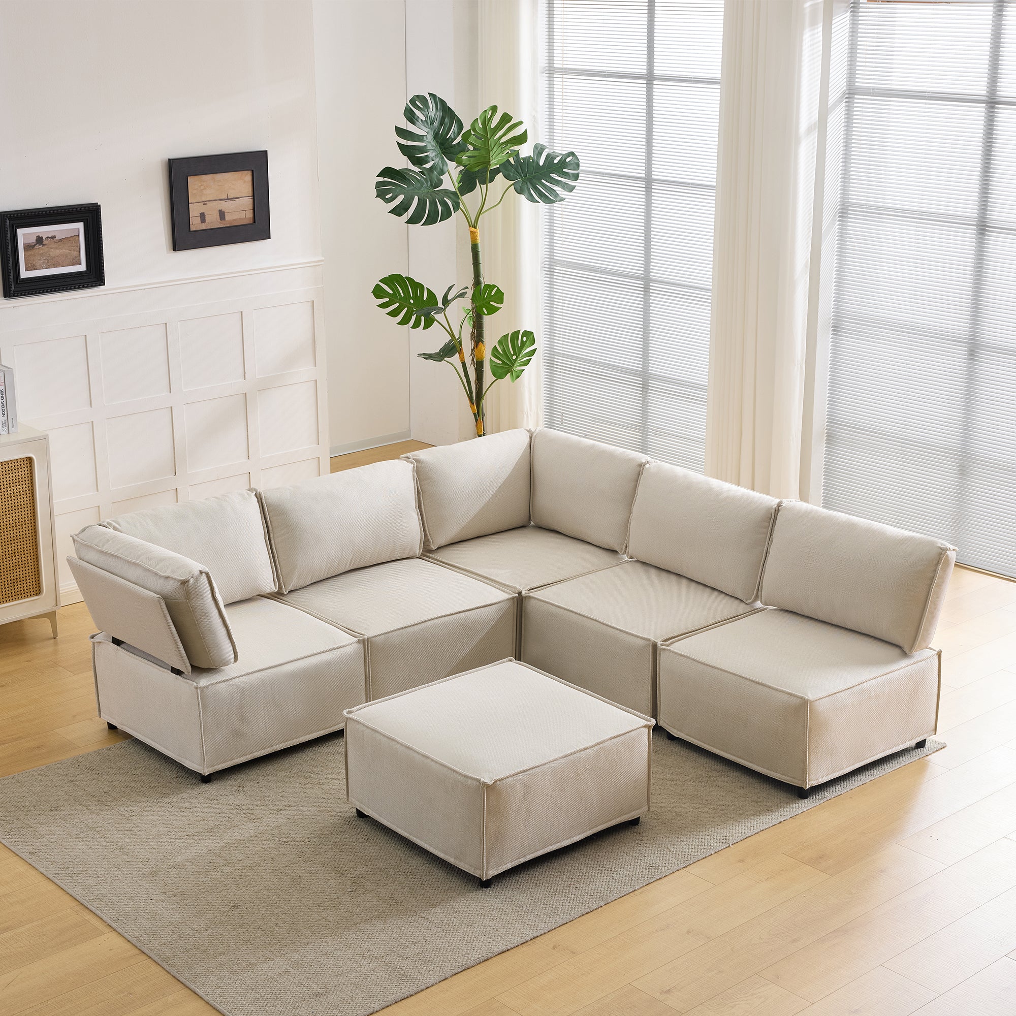 Beige sectional sofa with ottoman in a living room setting with a plant and framed pictures.